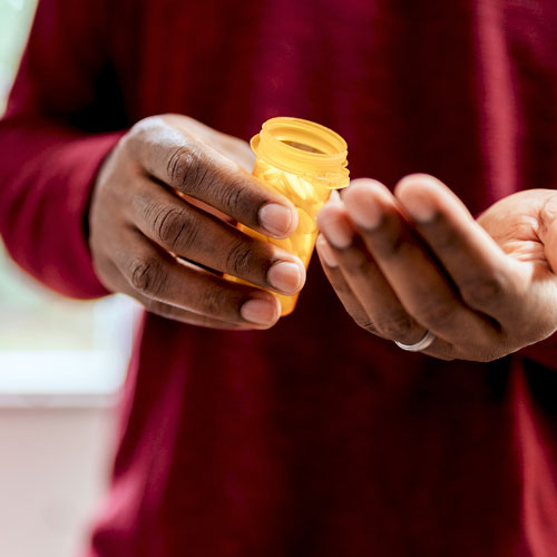Un hombre con una camiseta roja sostiene una botella de medicamento sin la tapa cuando está a punto de verter algunas pastillas en su mano.