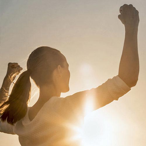 A woman in a white long-sleeve shirt, bathed in sunlight, raises her hands triumphantly after achieving her weight loss goals