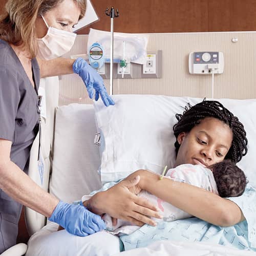 A high-risk pregnancy nurse stands at the bedside of a mother holding her newborn baby