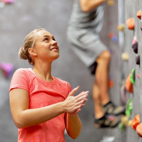 A young woman in a pink shirt rubs her hands together as she stands before a climbing wall