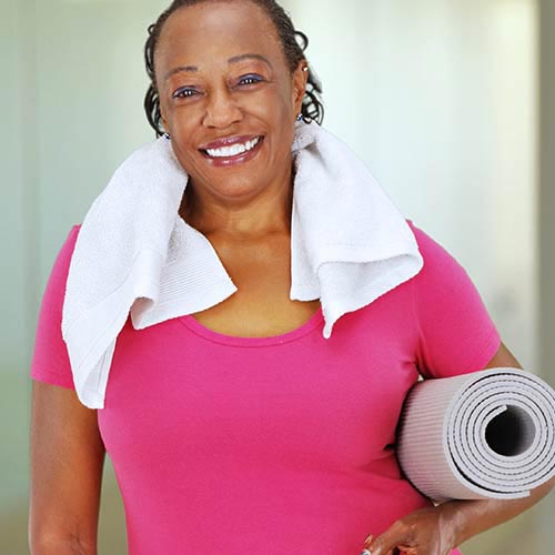 Una mujer sonriente con una camisa rosa con una toalla blanca alrededor del cuello y una alfombra de yoga enrollada debajo del brazo izquierdo