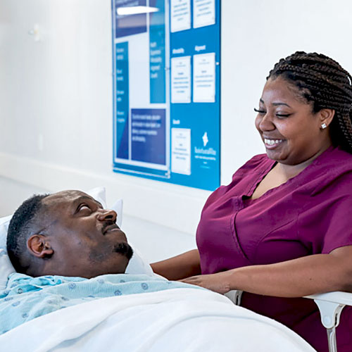 A nurse in magenta scrubs smiles while talking with a male patient lying on a bed on an inpatient floor