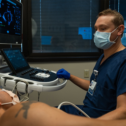 A sonographer sits at a machine while he performs a non-invasive cardiac procedure on a patient who is lying down